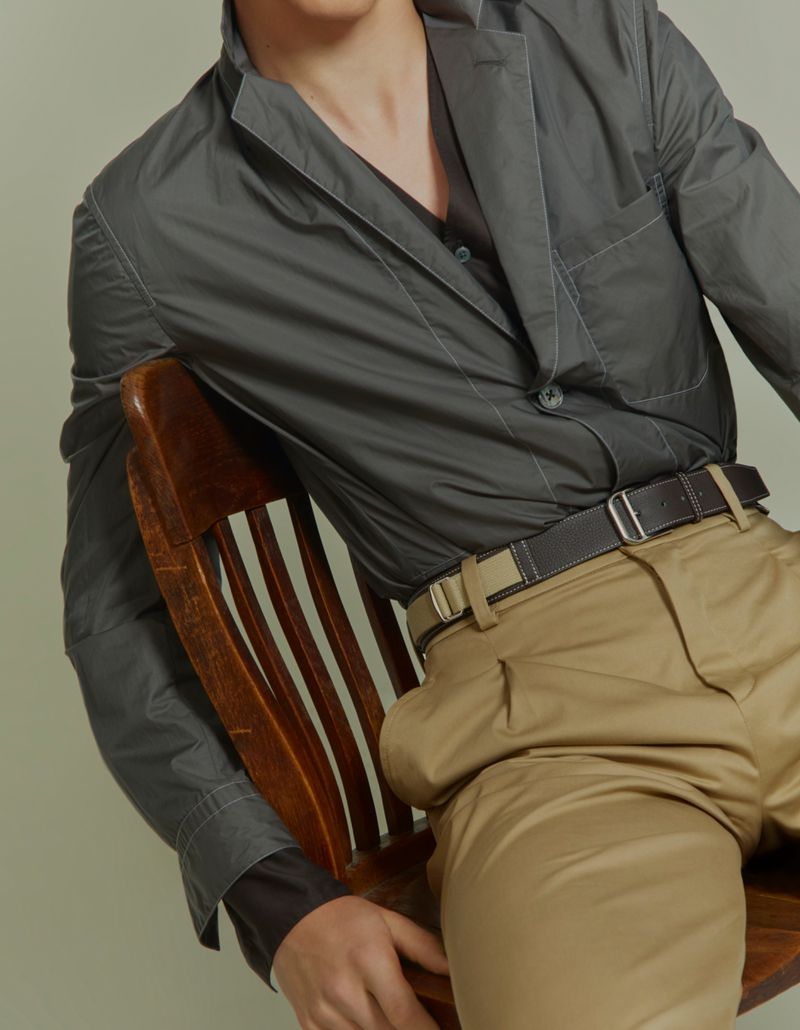 Model wearing a dark grey jacket and beige trousers seated on a wooden chair