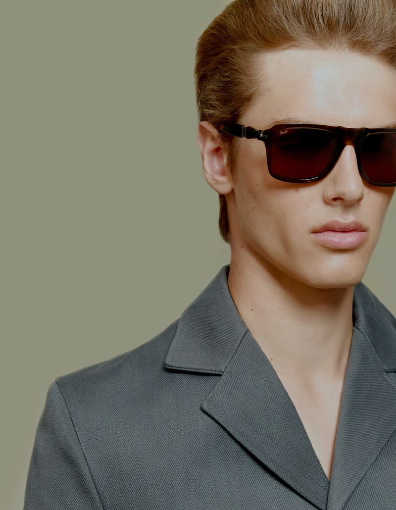 Model Esteban Monbaliu wearing dark rectangular sunglasses and a slate grey tailored jacket against a muted green background.