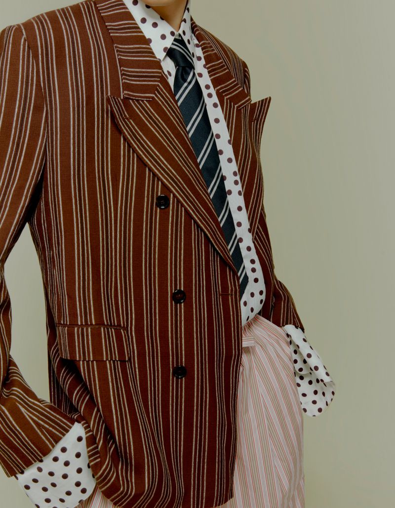 Close-up of a model wearing a brown pinstriped double-breasted blazer, polka-dot dress shirt, and striped tie against a plain backdrop.