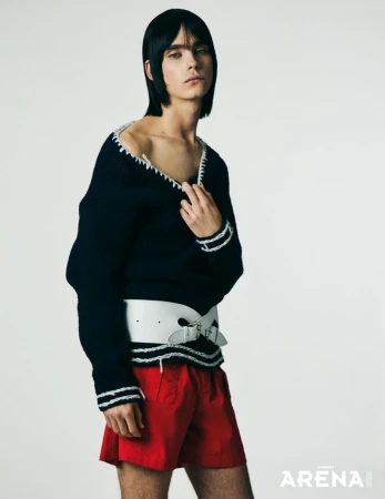 Model Frederic Bittner wearing an off-the-shoulder black knit sweater with a wide white belt and red shorts against a white background.