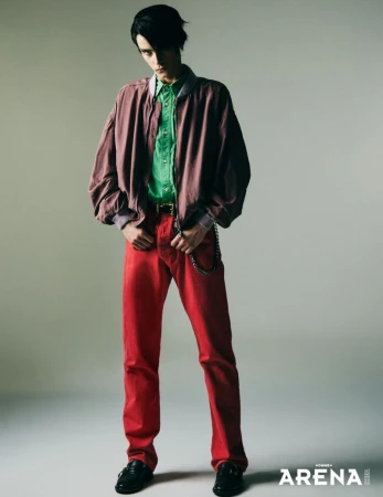 Male model wearing a mauve bomber jacket, green shirt, and red trousers standing against a pale grey studio background.