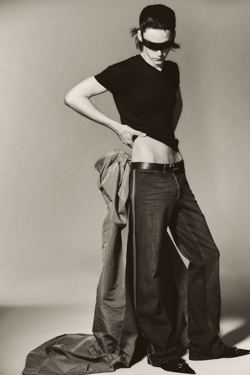 Model Frederic Bittner poses wearing a black t-shirt, dark wide-leg trousers, and a black visor against a seamless studio backdrop.