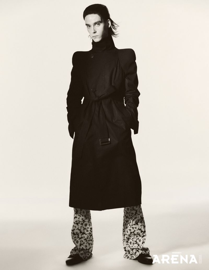 Model Frederic Bittner in a high-collared black trench coat with structured shoulders and printed trousers against a white backdrop.