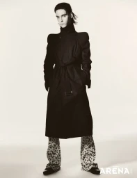 Model Frederic Bittner in a high-collared black trench coat with structured shoulders and printed trousers against a white backdrop.