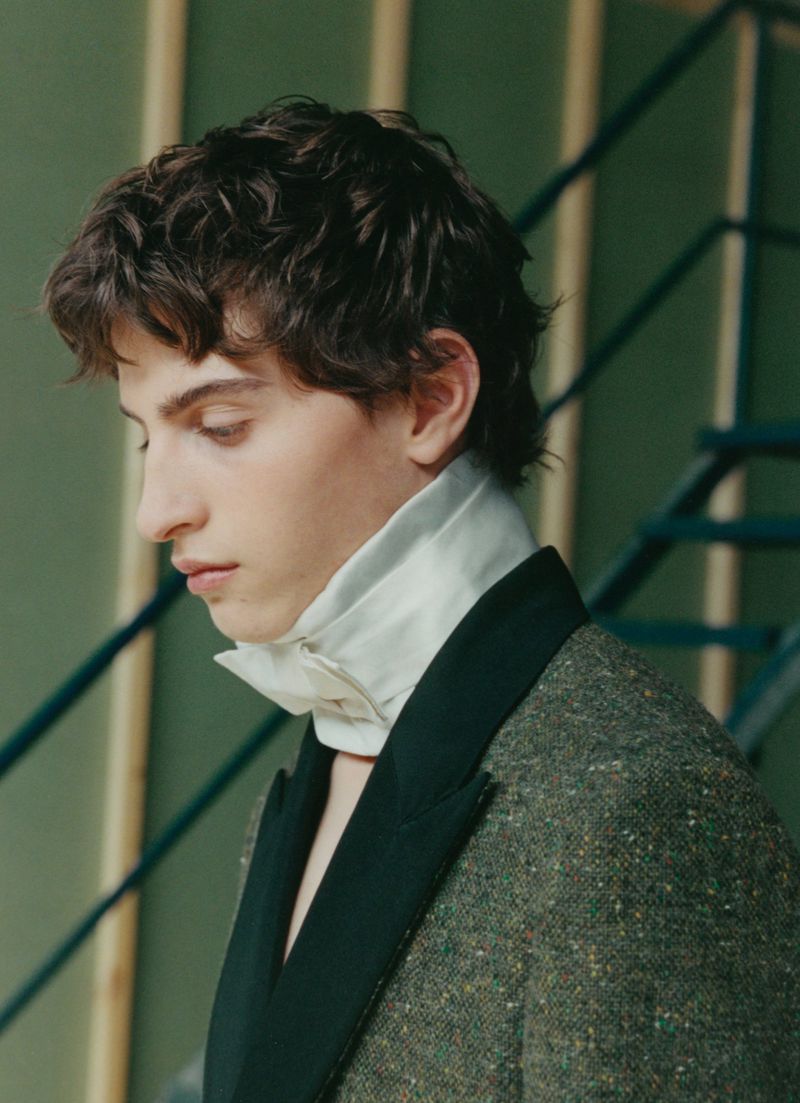 Profile portrait of a young man wearing a tweed blazer with black lapels and a high white collar shirt.
