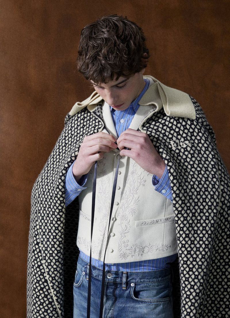 A young man looking down while tying the ribbons of an embroidered cream vest layered with a patterned Dior knit cape and blue jeans.