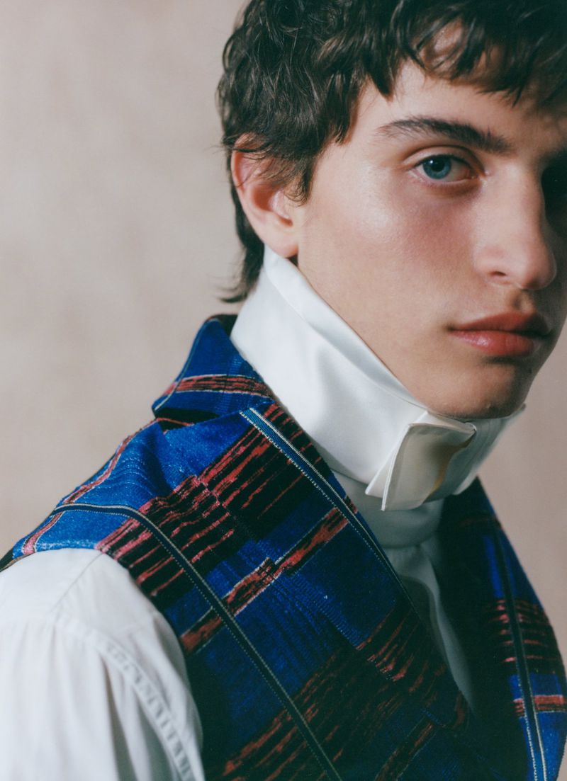 Close-up portrait of a young man wearing a high structured white collar and a blue patterned vest against a beige studio backdrop.