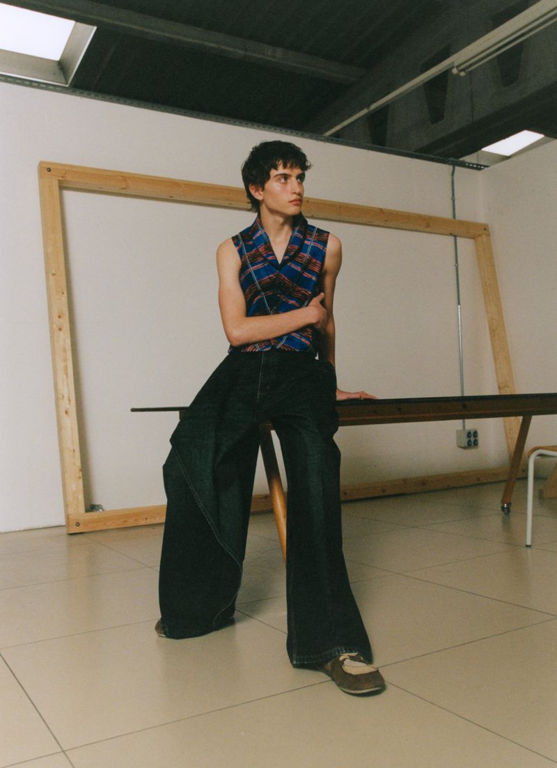 Model in a sleeveless plaid top and exaggerated wide-leg dark denim trousers leaning against a table in a studio.