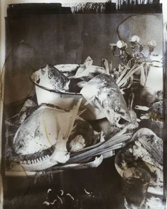 Monochrome vanitas still life with raw fish heads, an animal jawbone, leeks, and an ornate candelabra on a table.