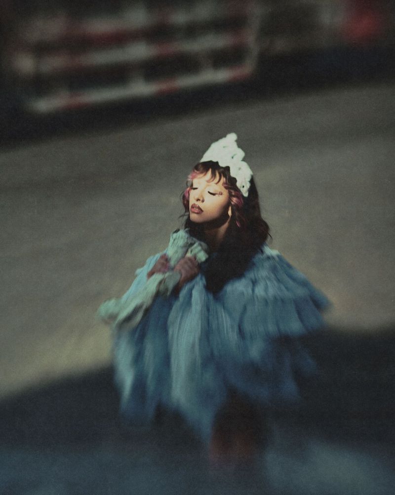 Melanie Martinez posing with eyes closed in a voluminous light blue ruffled gown and white lace headpiece in soft dim lighting.