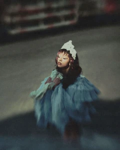 Melanie Martinez posing with eyes closed in a voluminous light blue ruffled gown and white lace headpiece in soft dim lighting.