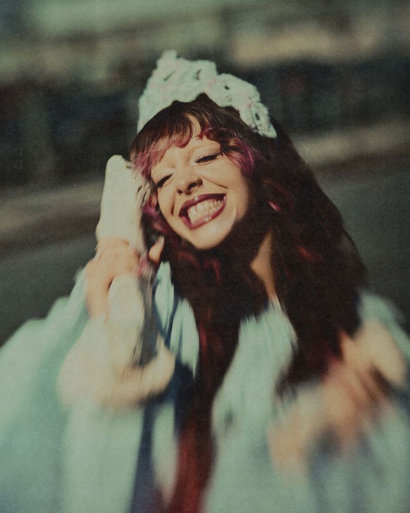 Melanie Martinez smiling widely in a light blue plush coat and pale lace crown for 1883 Magazine.