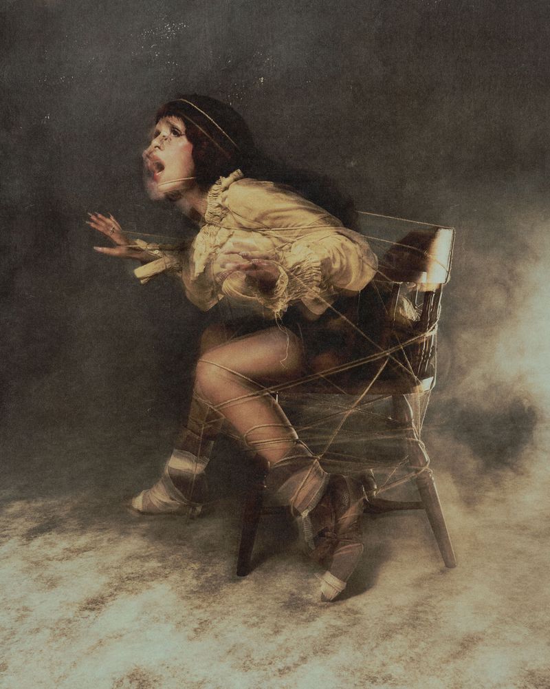 Melanie Martinez tied to a wooden chair with string, wearing a ruffled cream blouse in a smoky studio setting.