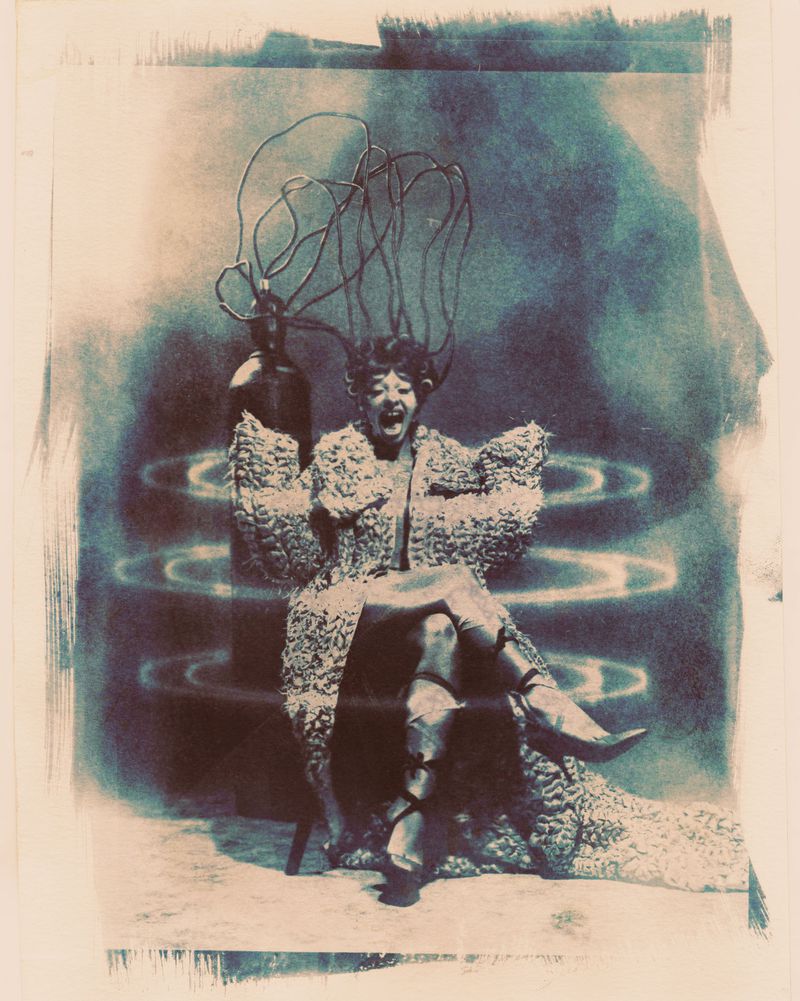 Melanie Martinez screaming while seated in an ornate coat with wires attached to her head and glowing light rings.