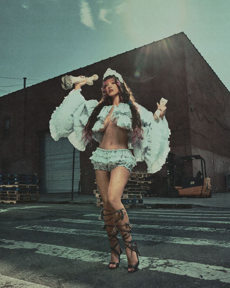Melanie Martinez posing in a ruffled light blue two-piece set and lace-up heels on an industrial street holding decorative props.