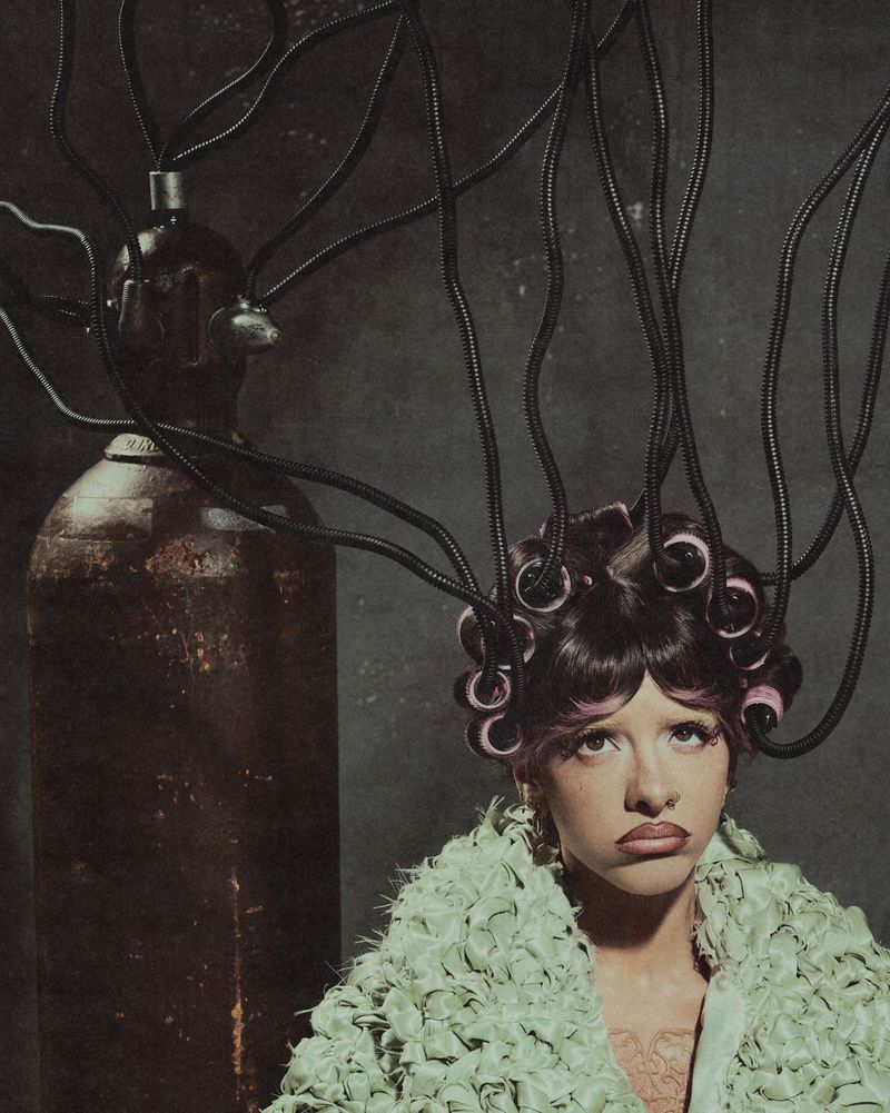 Melanie Martinez wearing a textured light green coat with hair curlers wired with black tubes to an industrial gas cylinder.