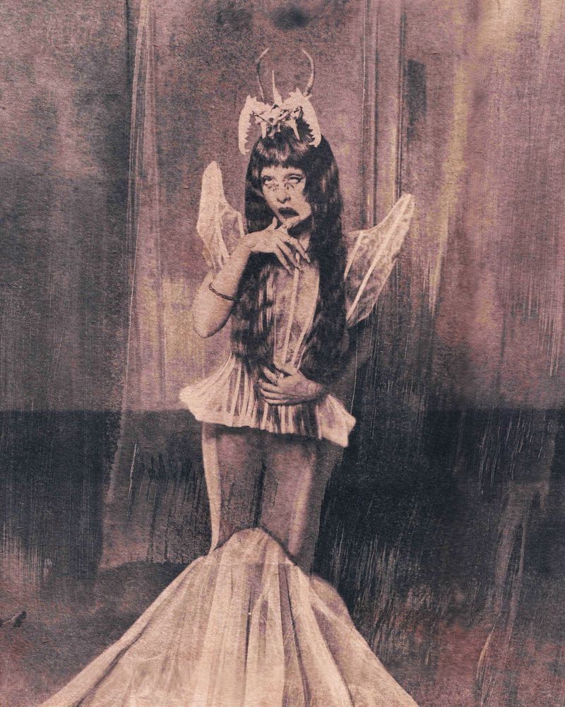 Melanie Martinez wearing a sculptural headpiece, translucent wings, and a mermaid gown in a moody textured editorial portrait.