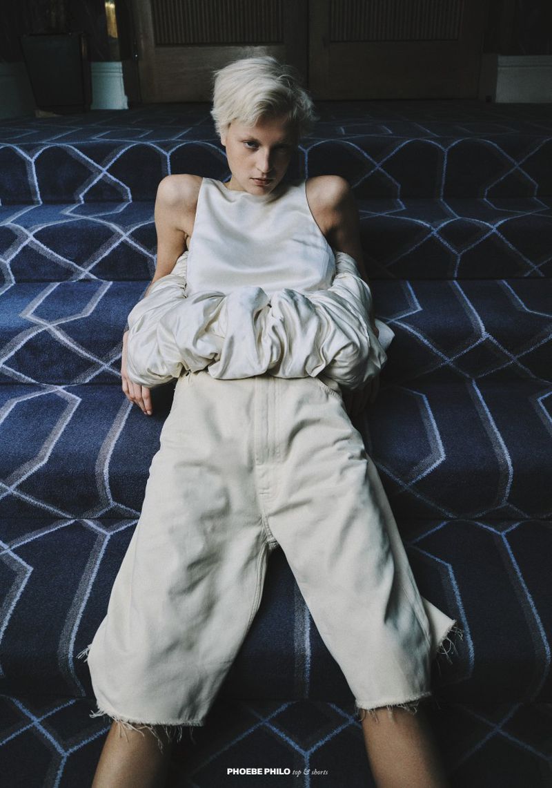 Model Michelle Laff with short blonde hair seated on blue patterned stairs wearing a white top and cream shorts for Puss Puss Magazine.