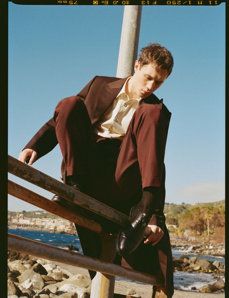 Male model wearing a brown tailored suit and light button-up shirt perched on a metal railing by a rocky coastline.