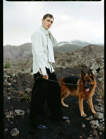 A male model wearing a white shirt and dark trousers standing outdoors on rocky ground holding a German Shepherd on a leash.