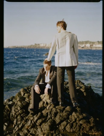 Two male models in tailored suits posing on dark coastal rocks with the deep blue ocean in the background.