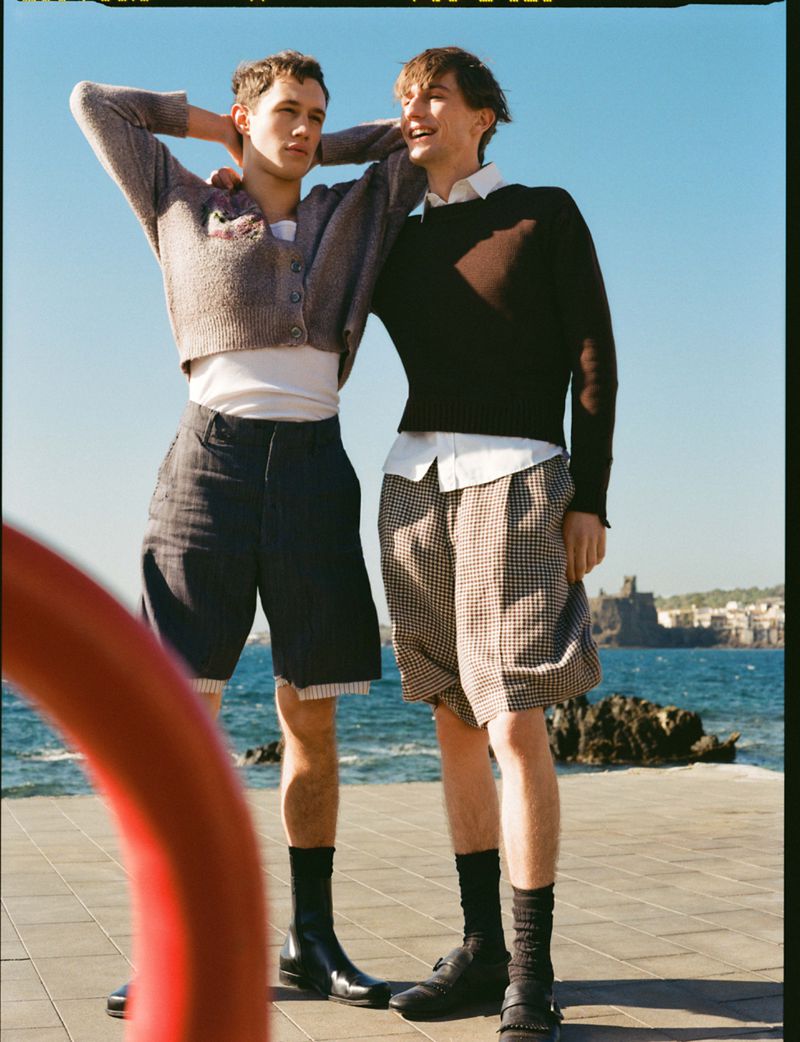 Two male models posing in tailored shorts, knit sweaters, and black shoes outdoors on a sunny paved promenade by the sea.