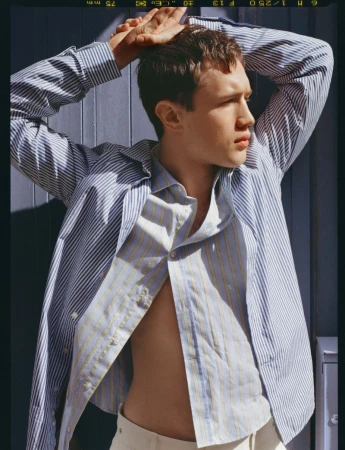 Fashion model in an unbuttoned blue and white striped shirt posing with hands behind head against a blue wall.