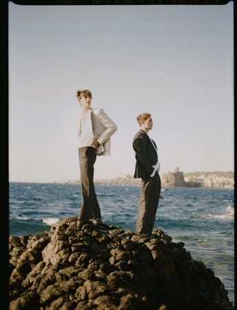 Two male models standing on a rocky outcrop by the ocean wearing tailored suits under clear sky