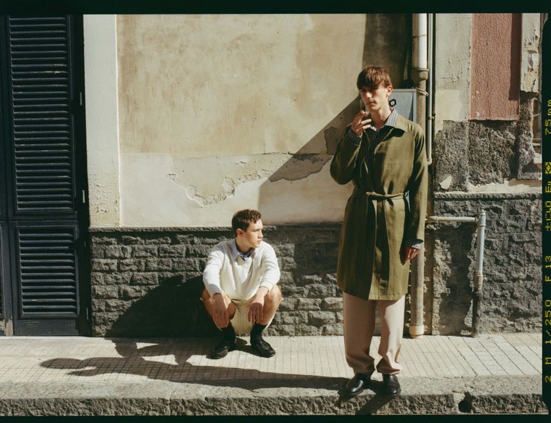 Two male models posing against a textured building wall in bright sunlight, one standing in an olive coat and one crouching.