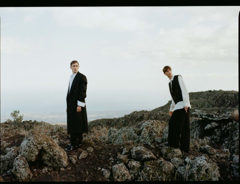 Two men in tailored black and white menswear standing on a rugged rocky hillside under an overcast sky.