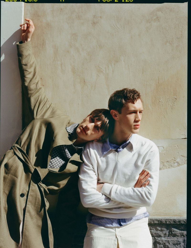 Two male models posing against a textured sunlit wall, one in an olive trench coat leaning on the other wearing a white sweater.