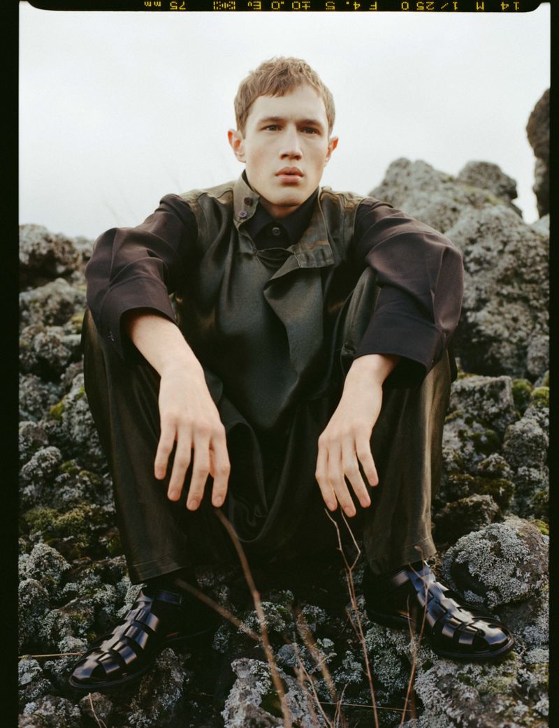 A young man crouches on mossy volcanic rocks wearing a dark layered tunic, trousers, and black fisherman sandals.