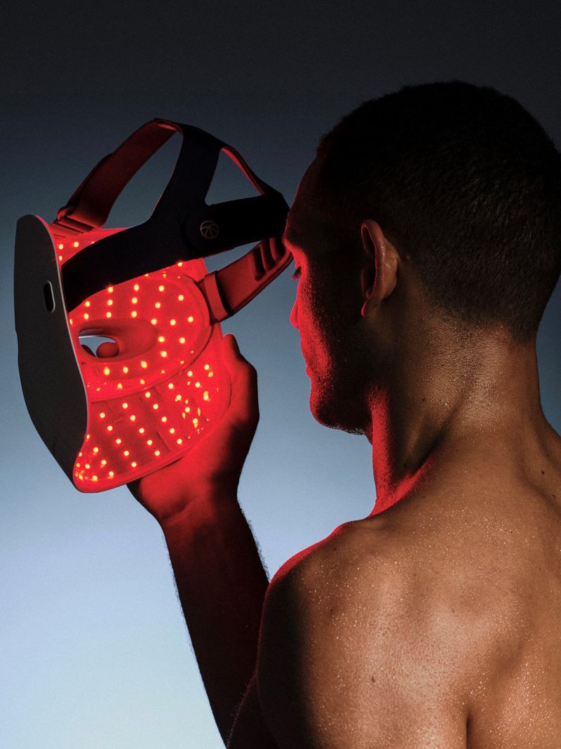 Shirtless man in profile holding an illuminated red LED light therapy face mask toward his face against a blue gradient backdrop.