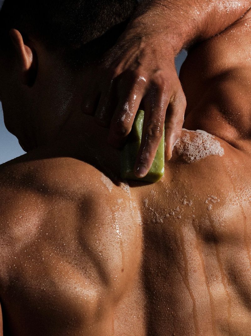 Close-up of a man's wet, muscular back as he scrubs his shoulder with a green bar of soap, creating white lather.