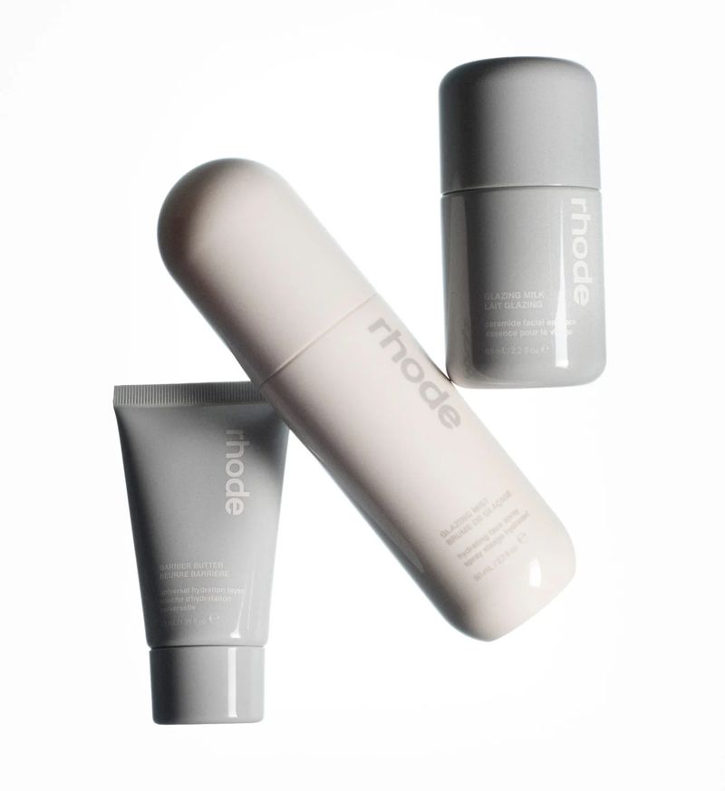 Three grey and white 'Rhode' brand skincare products are displayed on a white surface in an eye-level studio shot with copy on labels.
