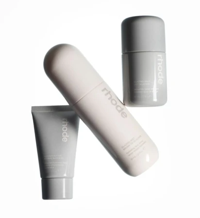 Three grey and white 'Rhode' brand skincare products are displayed on a white surface in an eye-level studio shot with copy on labels.