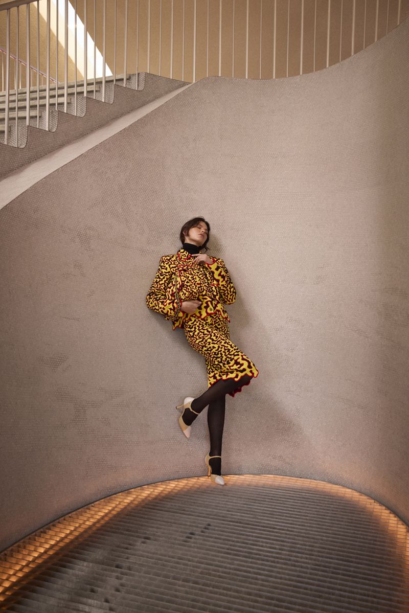 Model Georgia Fowler posing against a curved beige wall wearing a yellow patterned skirt suit, black tights, and white heels.