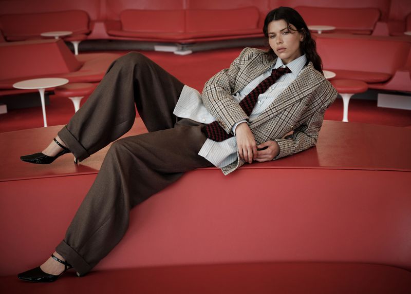 Model Georgia Fowler reclines on a curved red lounge bench wearing an oversized houndstooth blazer and wide-leg brown trousers.