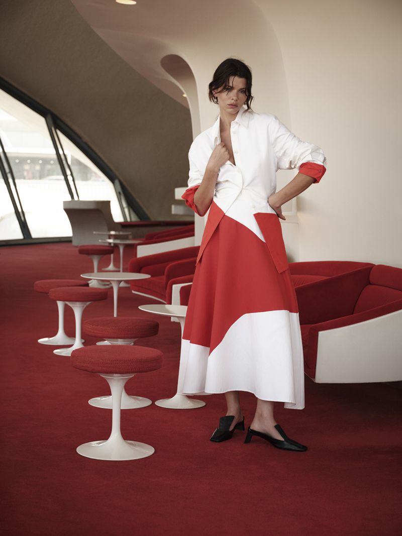 Model wearing a white and red color-blocked midi dress standing in a retro lounge with red carpet and white tulip stools.