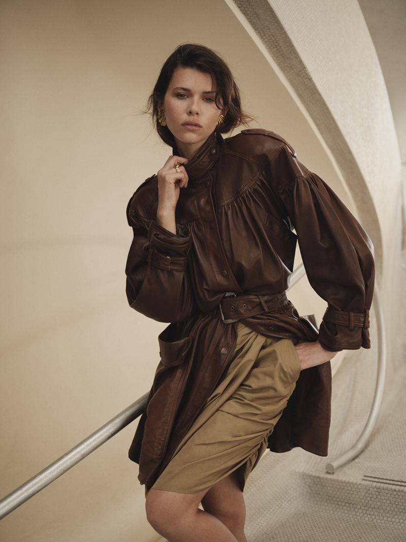 Model Georgia Fowler wearing an oversized brown leather trench coat and beige skirt on a modern curved staircase.