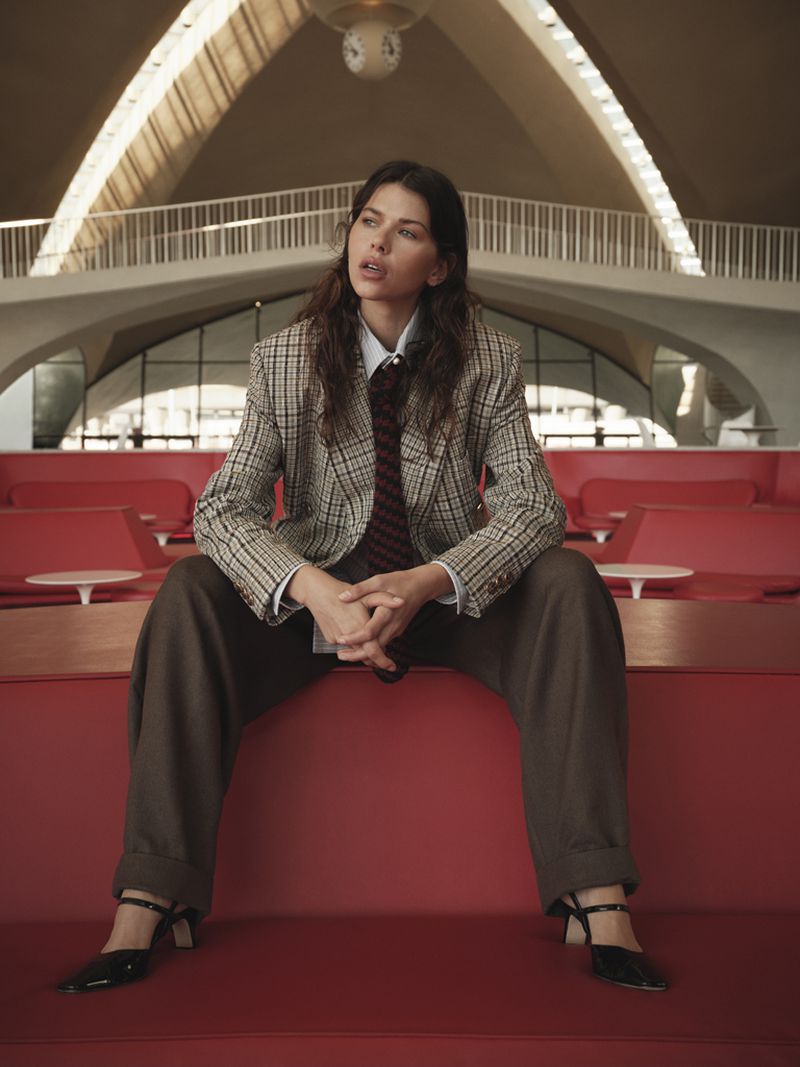 Model Georgia Fowler seated in a plaid blazer, necktie, wide-leg brown trousers, and low heels against a red architectural lounge.