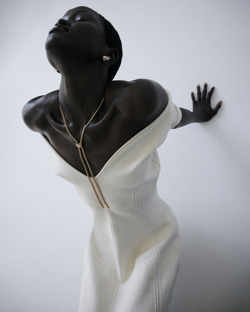 Model Anyiel Majok in an off-the-shoulder white dress with a gold lariat necklace leaning against a white studio wall.