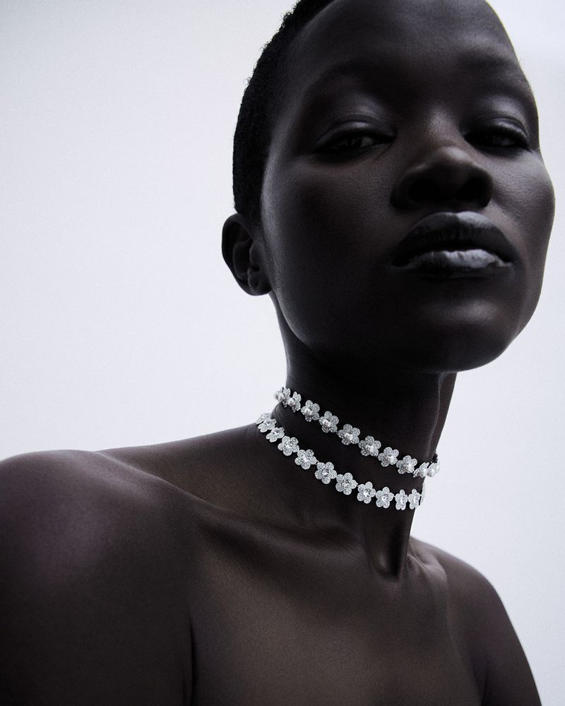 Model Anyiel Majok poses with bare shoulders wearing a sparkling floral choker necklace against a minimal grey studio backdrop.