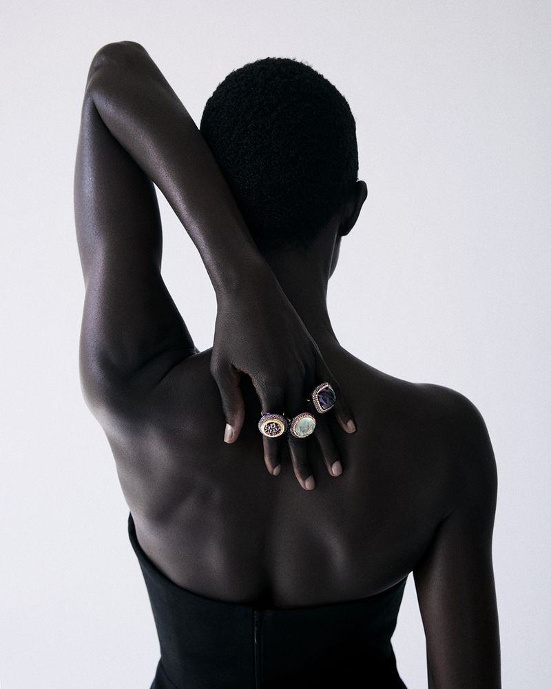 Model Anyiel Majok viewed from behind displaying gemstone cocktail rings with hand on spine against a white seamless backdrop.