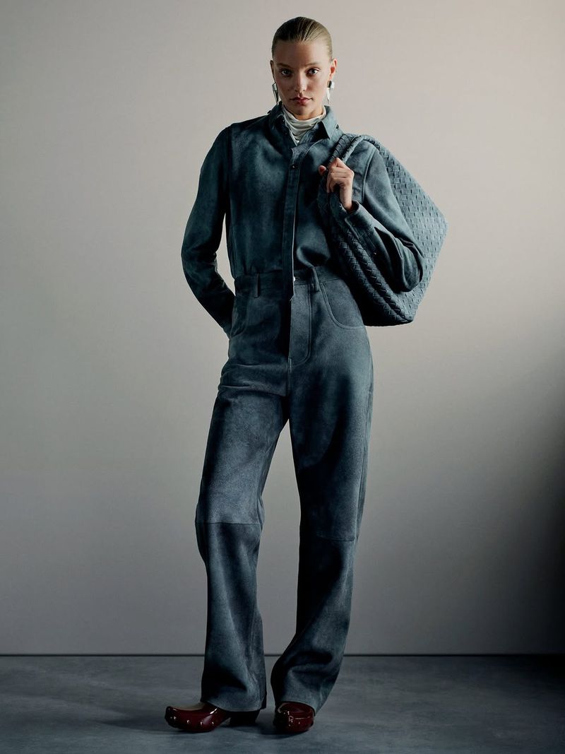 Model standing in a matching grey-blue shirt and trousers set holding a large woven leather shoulder bag against a grey background.