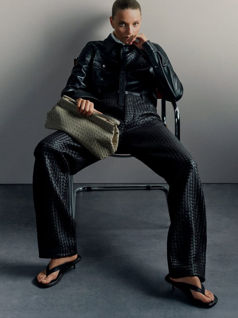 Model Taylor D seated in a chrome chair wearing a black leather shirt, woven trousers, and heeled sandals with an oversized clutch.