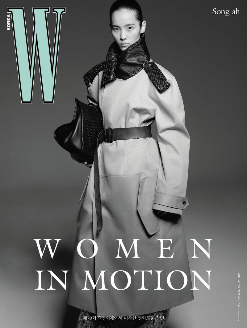 Model wearing a tan belted coat and dark woven scarf holding a black leather handbag against a grey backdrop for W Korea