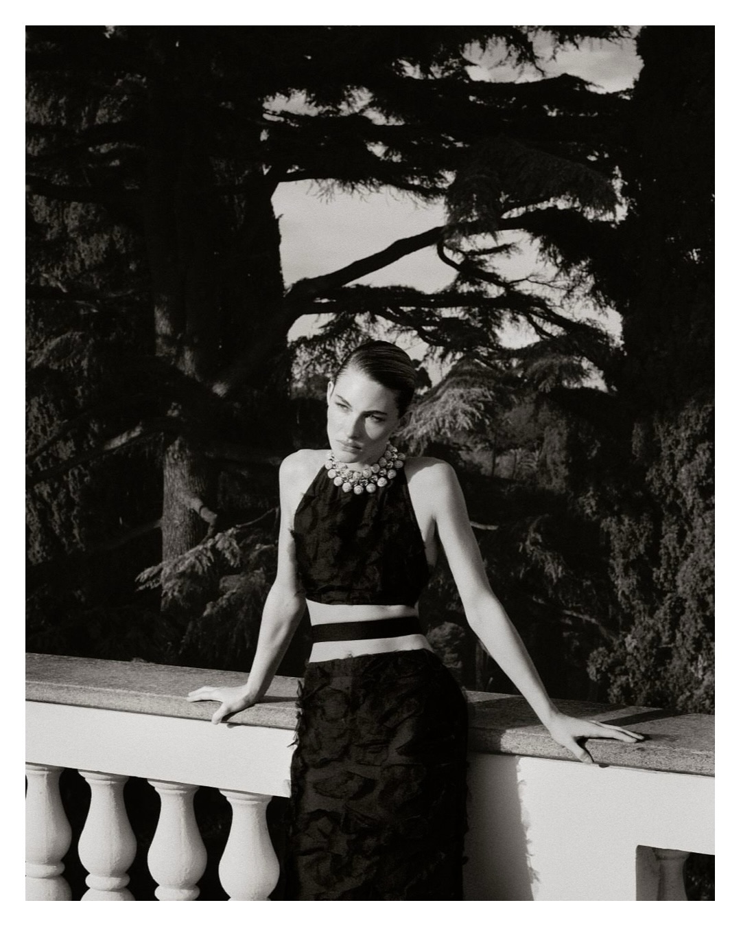 Grace Elizabeth leaning against a stone balustrade wearing a black textured two-piece outfit and statement pearl necklace in monochrome.