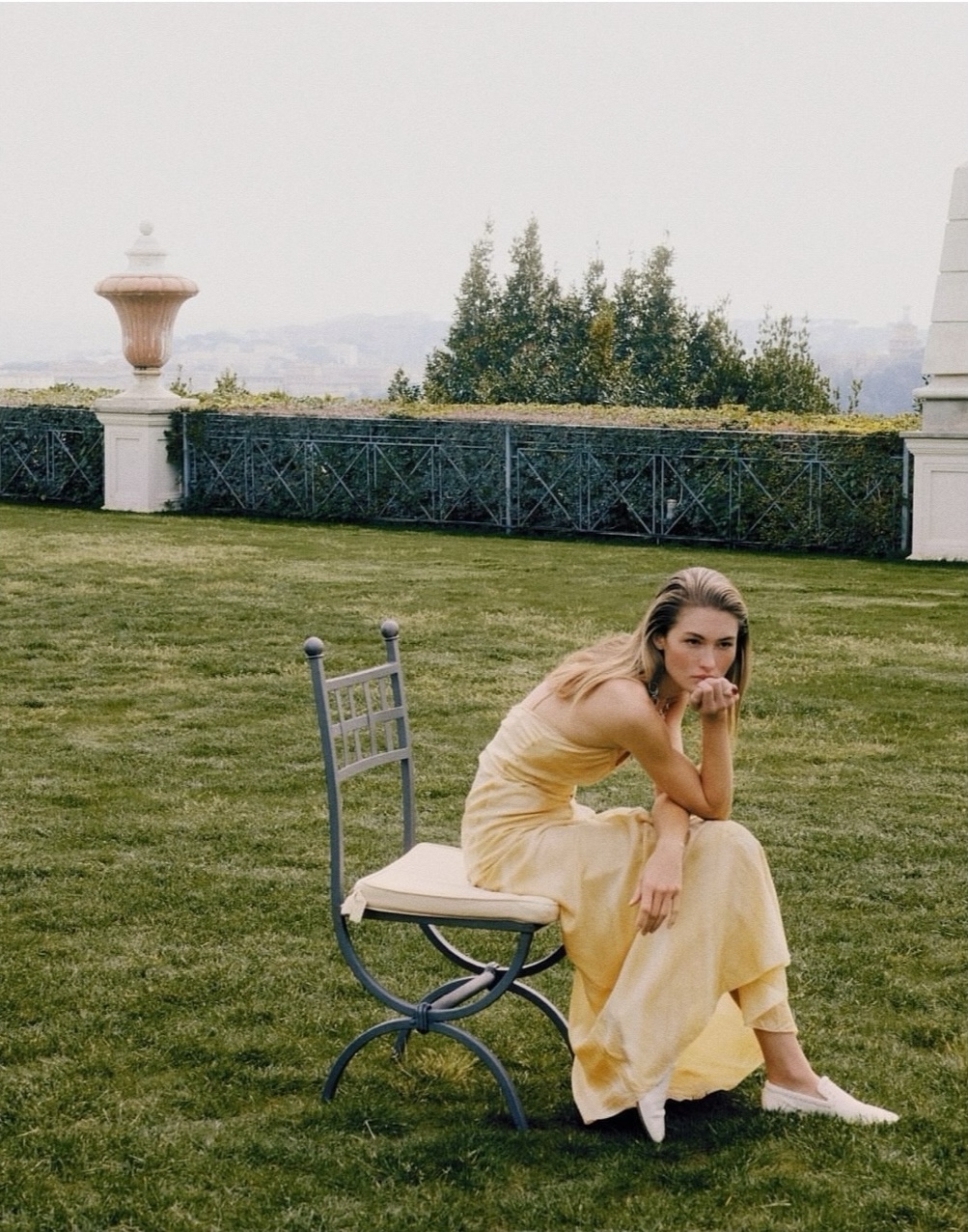Model Grace Elizabeth in a pale yellow strapless dress sits on a metal garden chair across an expansive manicured lawn.