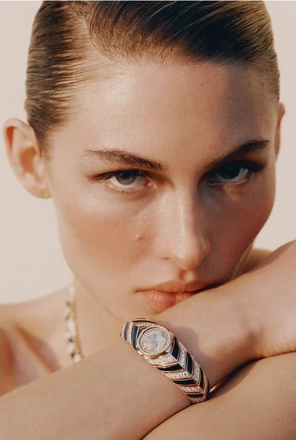 Close-up portrait of model Grace Elizabeth resting her chin near a luxury diamond and black chevron wristwatch against a pale background.